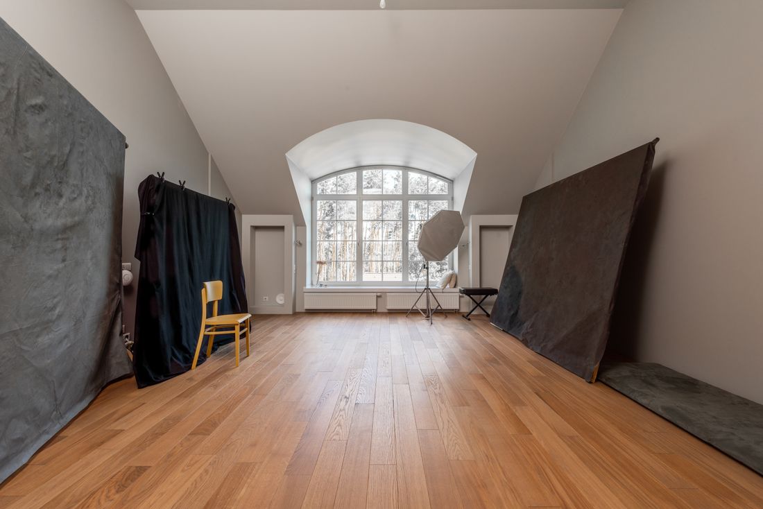 An empty photography studio with wood floors, dark fabric backdrops, and an arched window letting in daylight