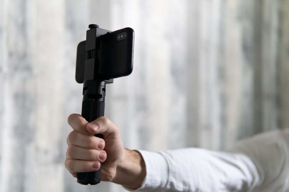 A hand holding a smartphone mounted on a small handheld gimbal, framed close against a blurred wall