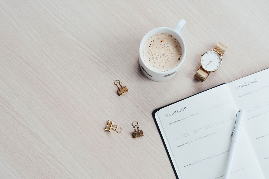 An open planner and pen laid out on a desk beside a coffee cup and wristwatch, seen from above