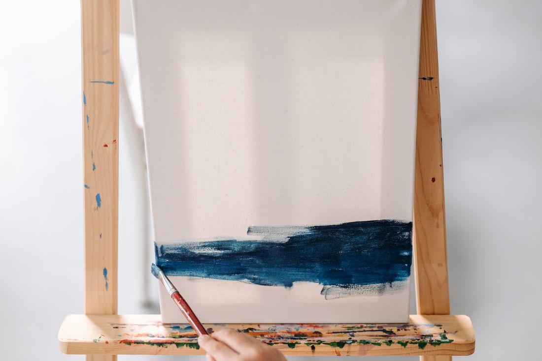 An unfinished blue wash on paper clipped to a wooden easel with a brush resting below