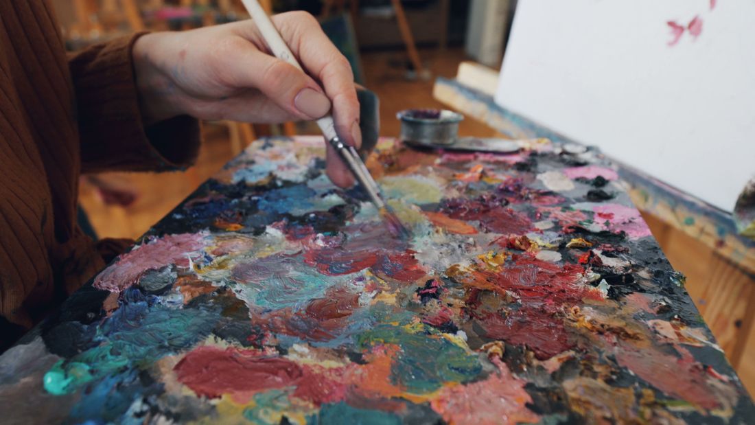 A hand mixing thick paint with a palette knife across a heavily used palette