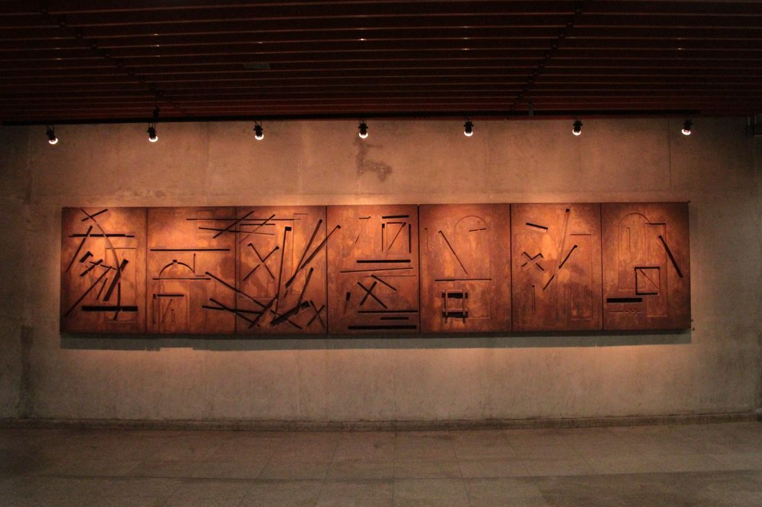 A long horizontal work lit against a dark wall in a dim exhibition room