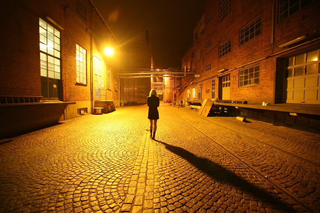 A woman walking alone down a lamplit cobblestone alley at night, the walk between one gallery opening and the next