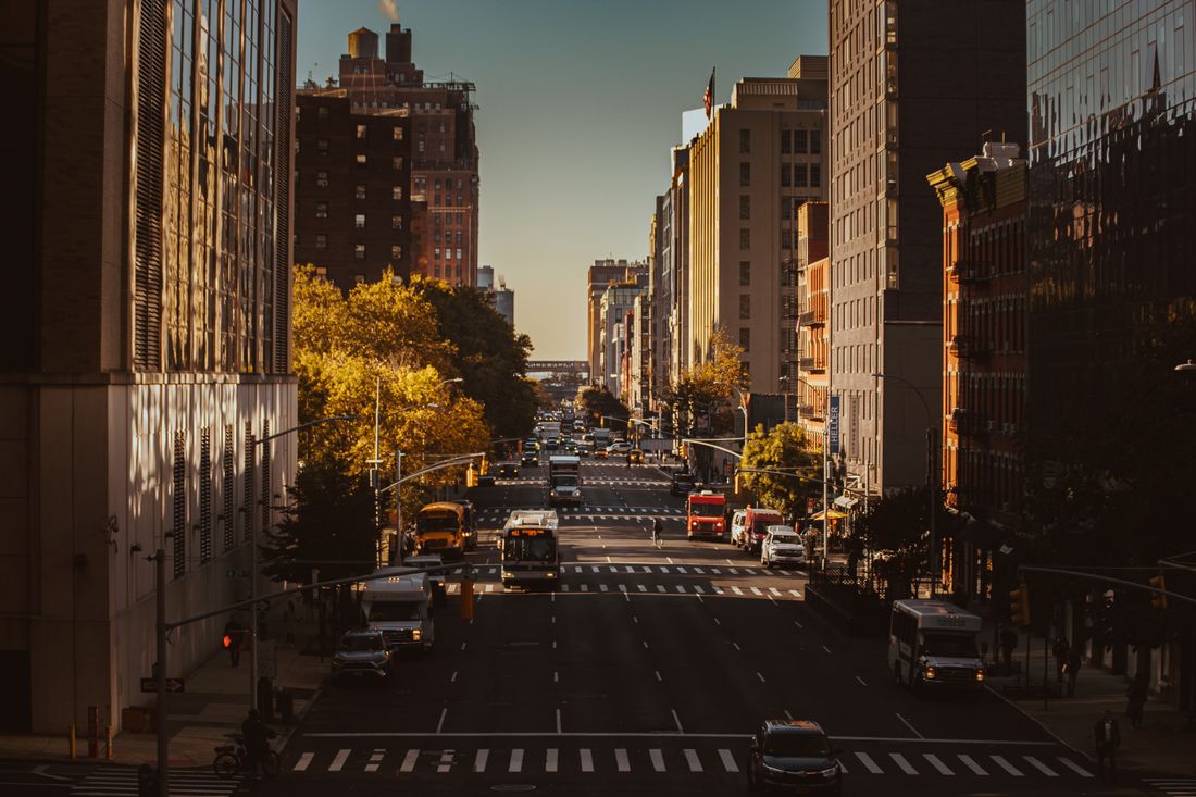 An elevated view down a wide Manhattan avenue at golden hour, buses and cars in traffic between tall buildings.