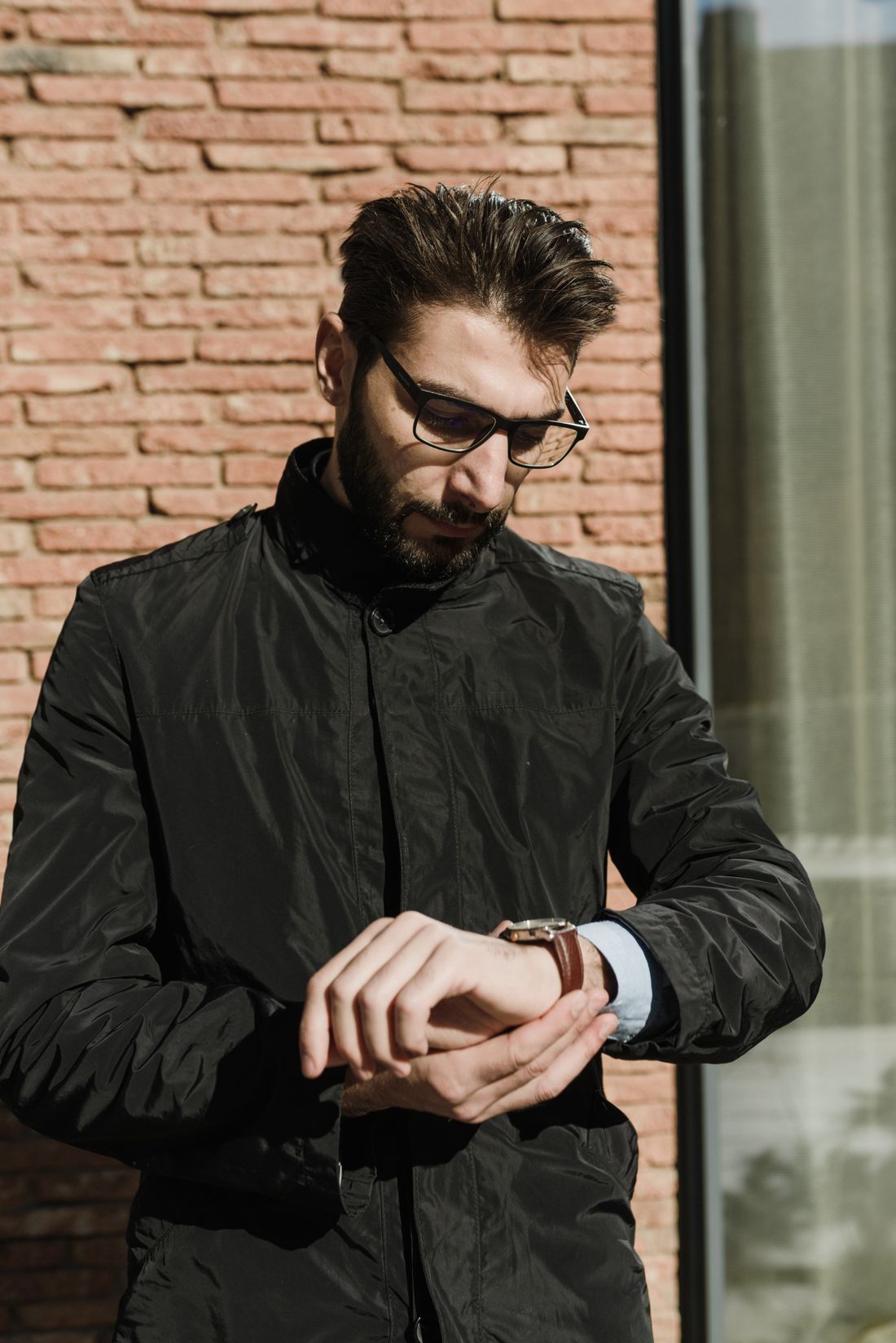 A man in a dark jacket standing outdoors against a brick wall, checking the time on his wristwatch.