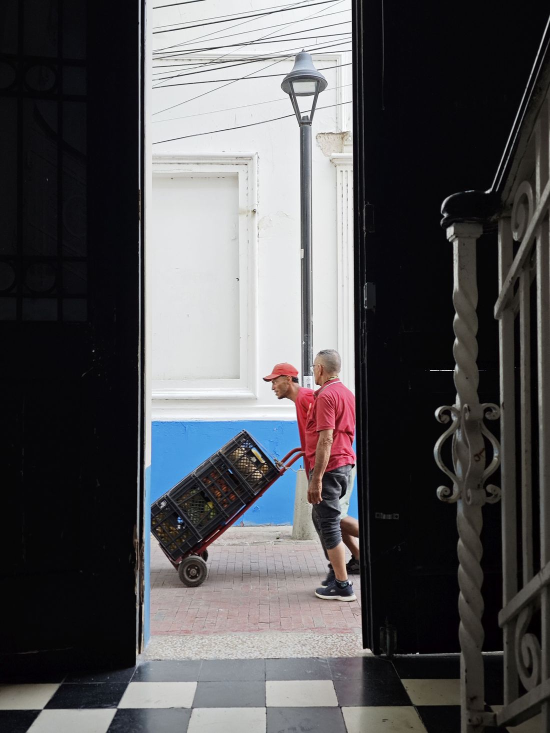 Two people seen through an open doorway pushing a hand cart loaded with a crate across a paved courtyard.