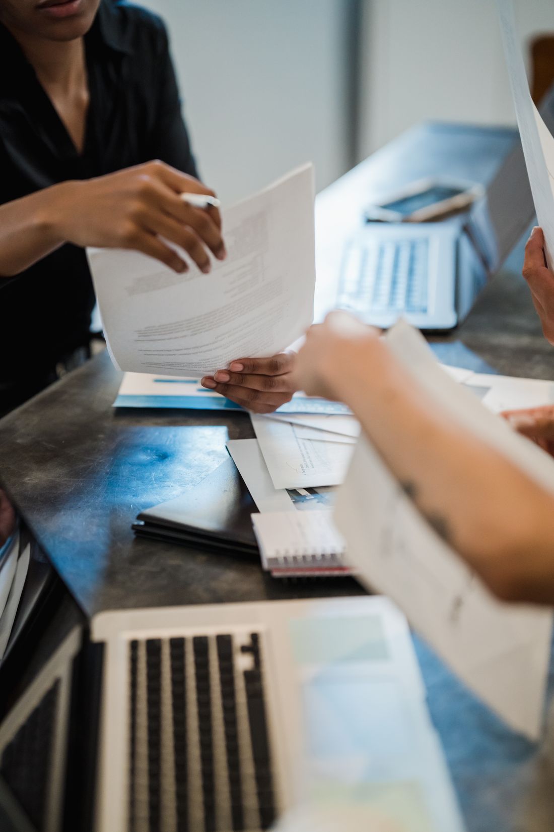 Two people exchanging a printed document across a desk with laptops and a calculator nearby.