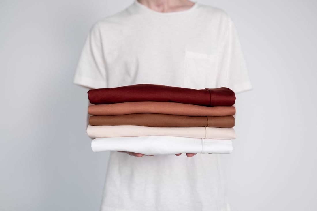 A close-up of a person holding a stack of neatly folded neutral-colored shirts
