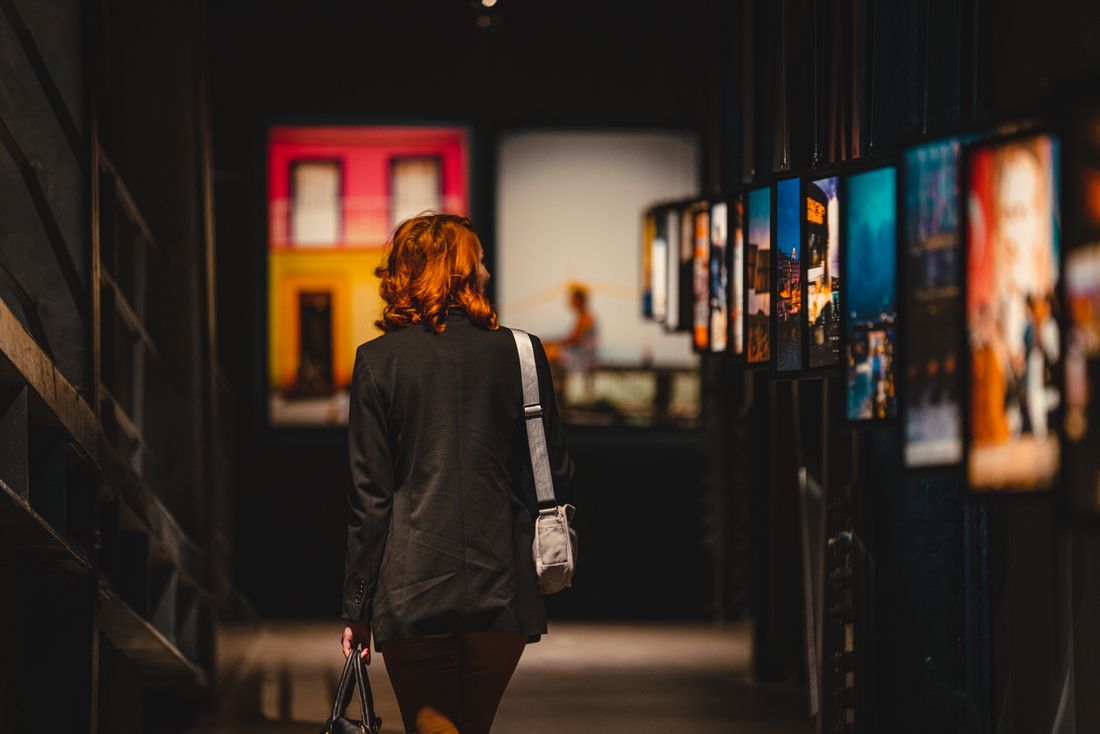 A visitor seen from behind, bag over one shoulder, walking slowly past a wall of framed photography in a gallery corridor.