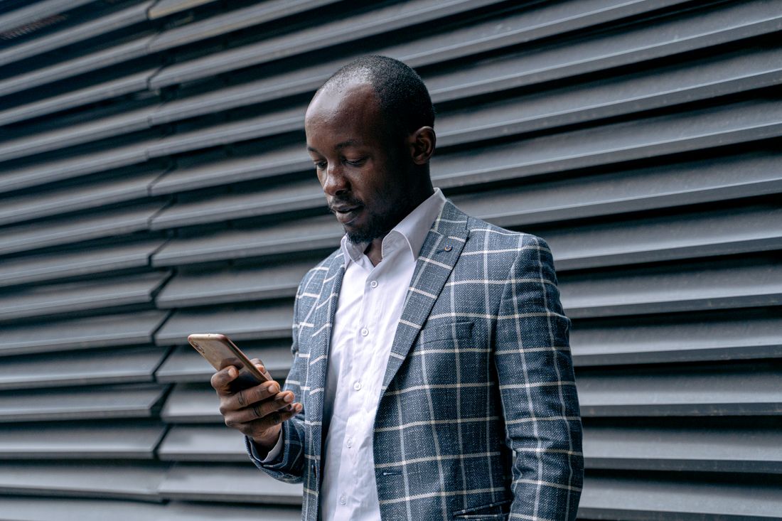 A man in a jacket stands against a plain slatted wall, looking down at his phone, seen from the side