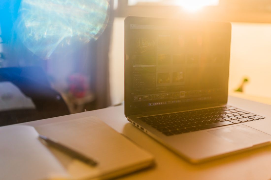An open laptop on a desk shows a blurred grid of images on screen, warm daylight flaring in, a closed notebook and pen beside it