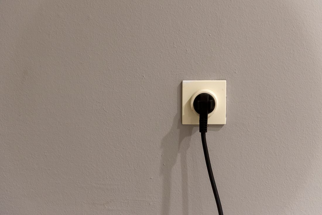 Close-up of a wall power outlet with a black cord plugged in, against a plain grey wall.