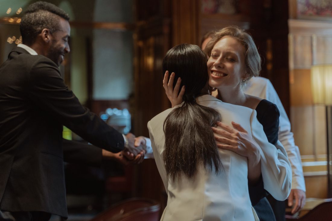 Two people greeting each other warmly indoors, with a third person shaking hands nearby