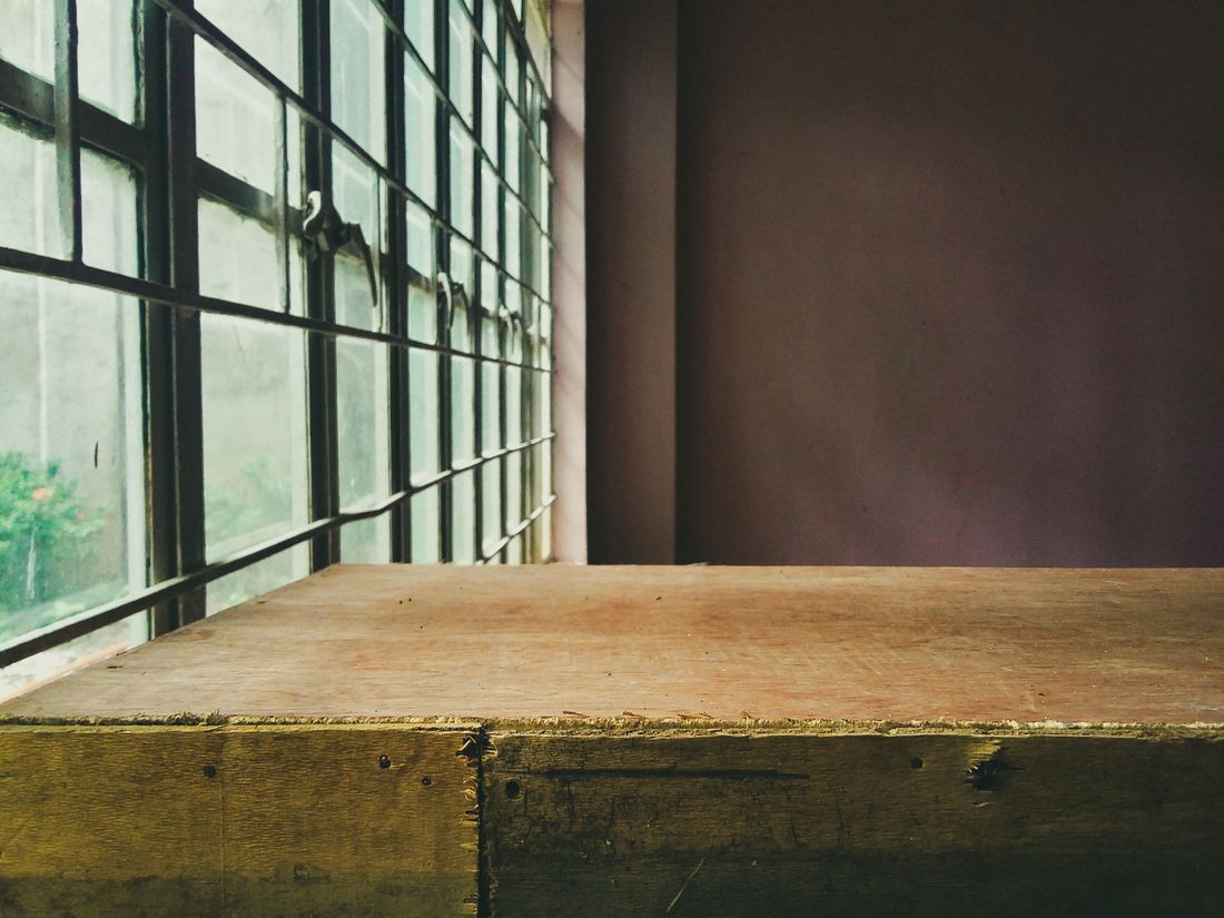 A cleared plywood work table beside a large industrial window, with a plain wall behind it and an empty room.