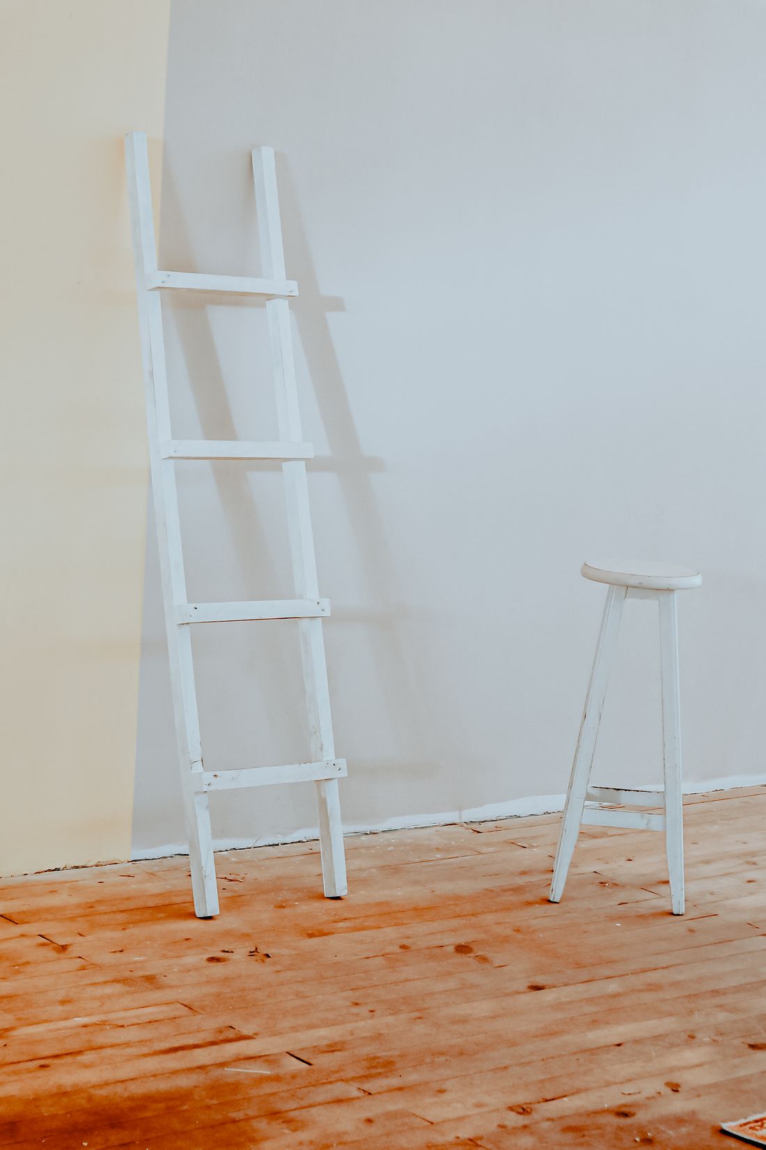 A white wooden ladder leaning against a plain wall next to a stool, in a bright room with a bare wood floor.