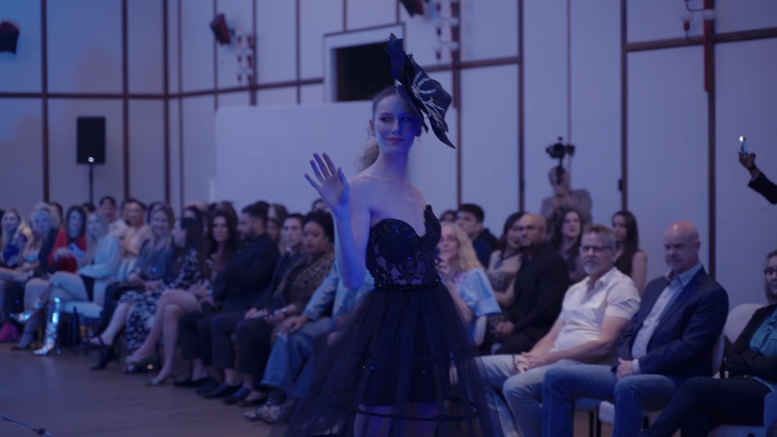 A model walks a runway under blue stage light while a seated audience watches and photographs the look from both sides.