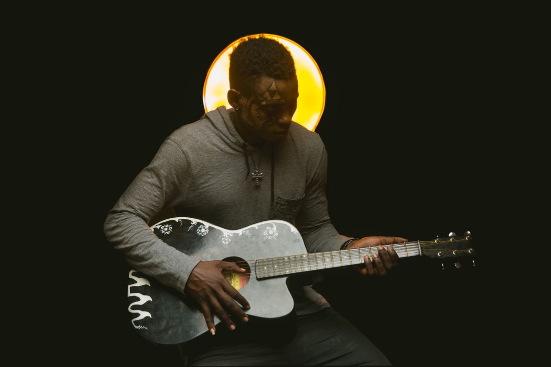 A musician with face paint plays acoustic guitar under a warm spotlight against a dark background
