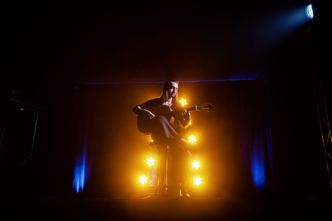 Musician playing acoustic guitar on stage, illuminated by dramatic yellow and blue lights.