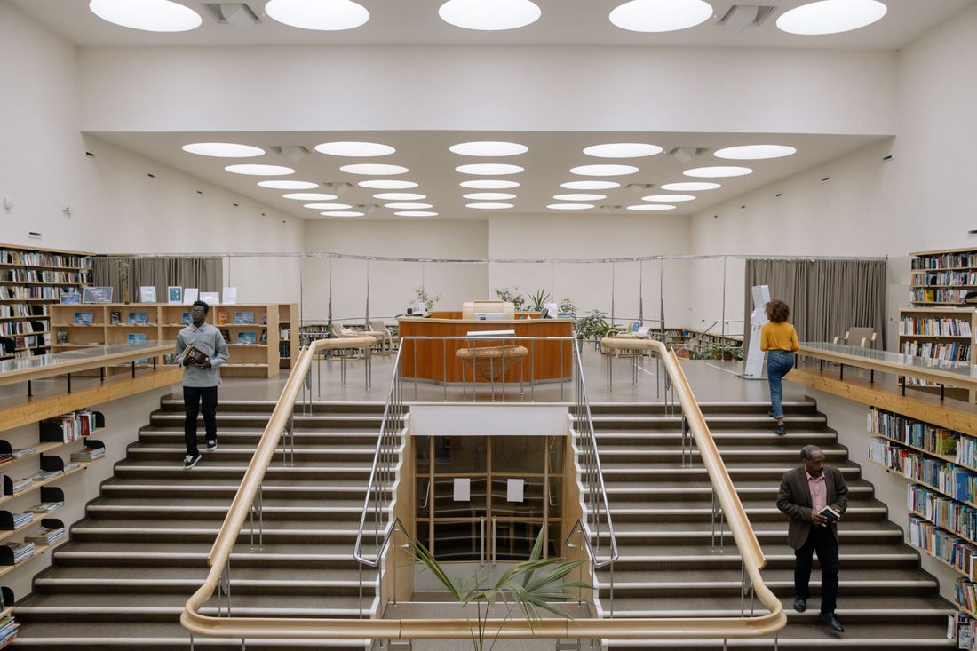 A wide institutional interior with a skylight ceiling, bookshelves lining both sides, and a few small figures on the stairs and upper level