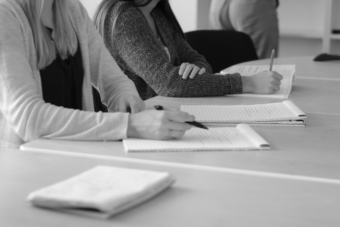 Close view of two people seated side by side at a shared table, writing quietly in notebooks with faces out of frame