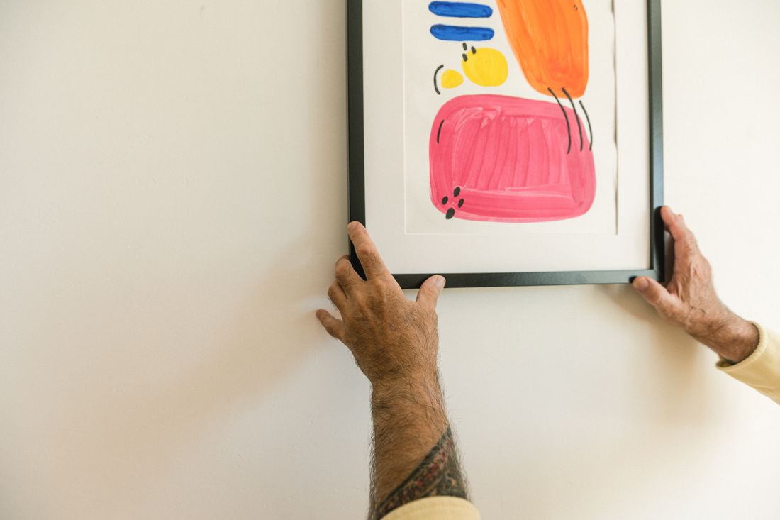 A pair of hands hanging a colorful abstract framed artwork on a white wall