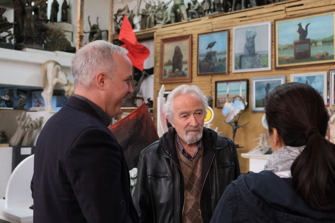 Three adults standing and talking together in a studio filled with sculptures and framed paintings