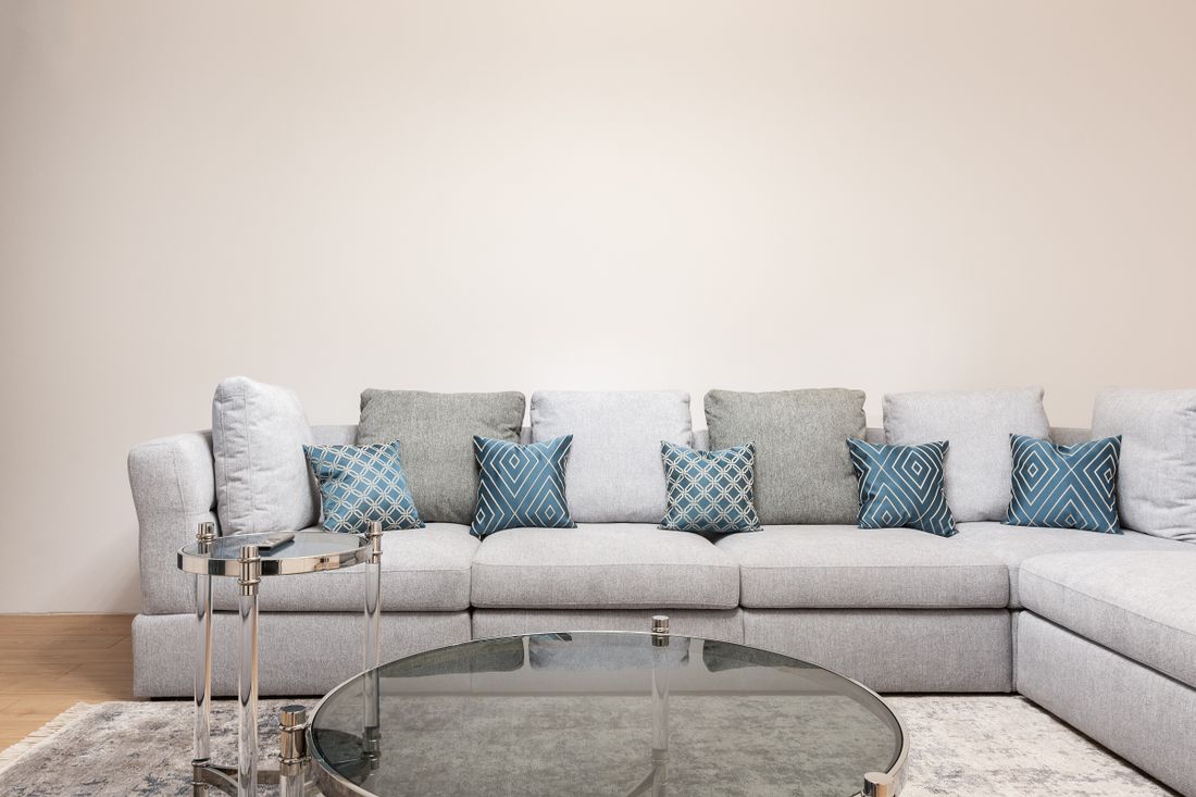 A bright, furnished living room with a light-colored sectional sofa and a coffee table in front of a plain, bare wall with nothing hung on it.