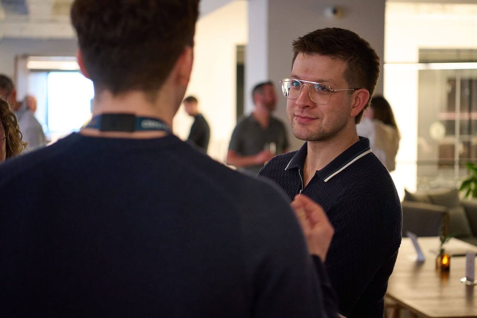 Two men in conversation at an indoor gallery event, one facing the camera in glasses, the other seen from behind wearing a lanyard.