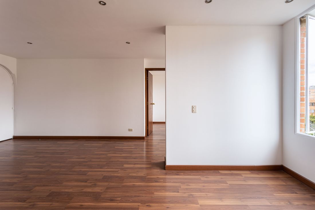 An empty white-walled room with polished wood flooring, an open interior doorway, and soft daylight, with no artwork hung.