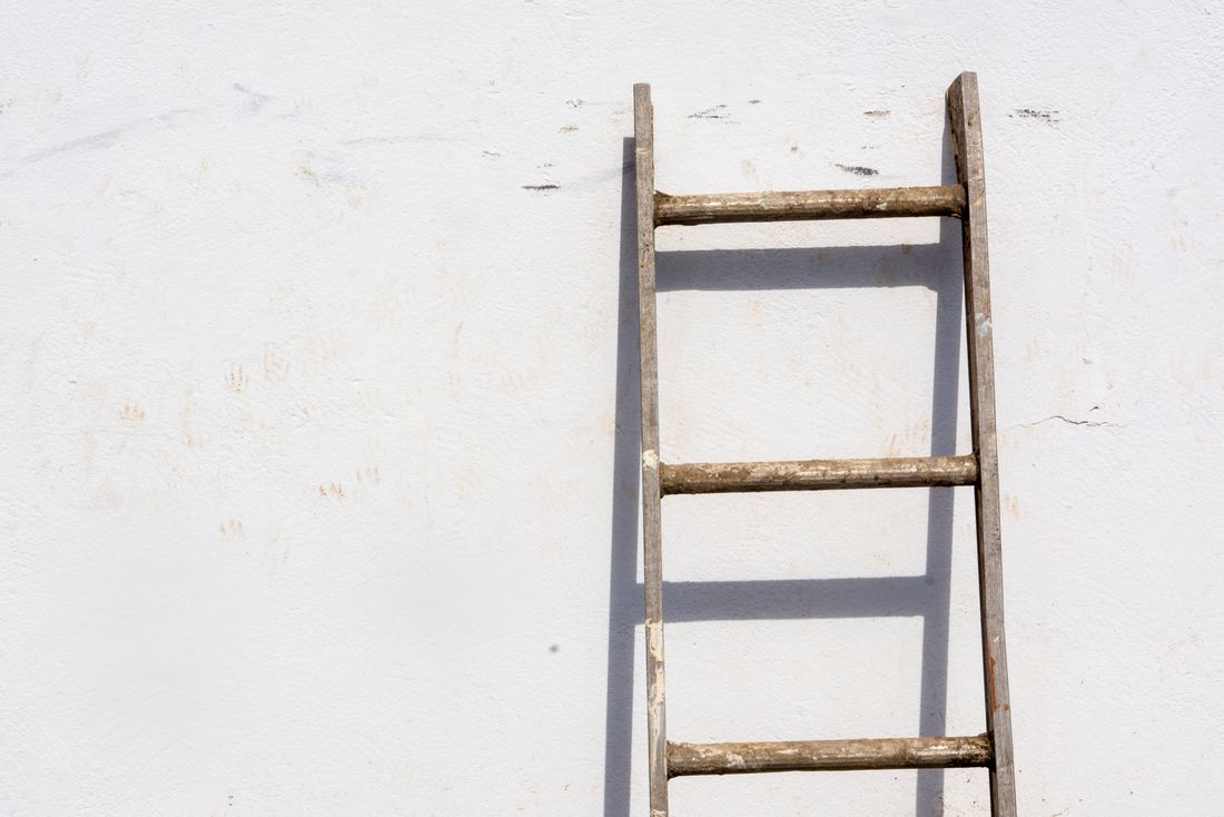 A weathered wooden ladder leaning against a plain white wall, off-center with open wall space around it.