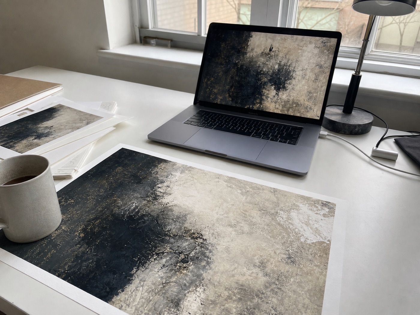Casual desk view of an artwork print beside a laptop, proof papers, a mug, and a USB cable