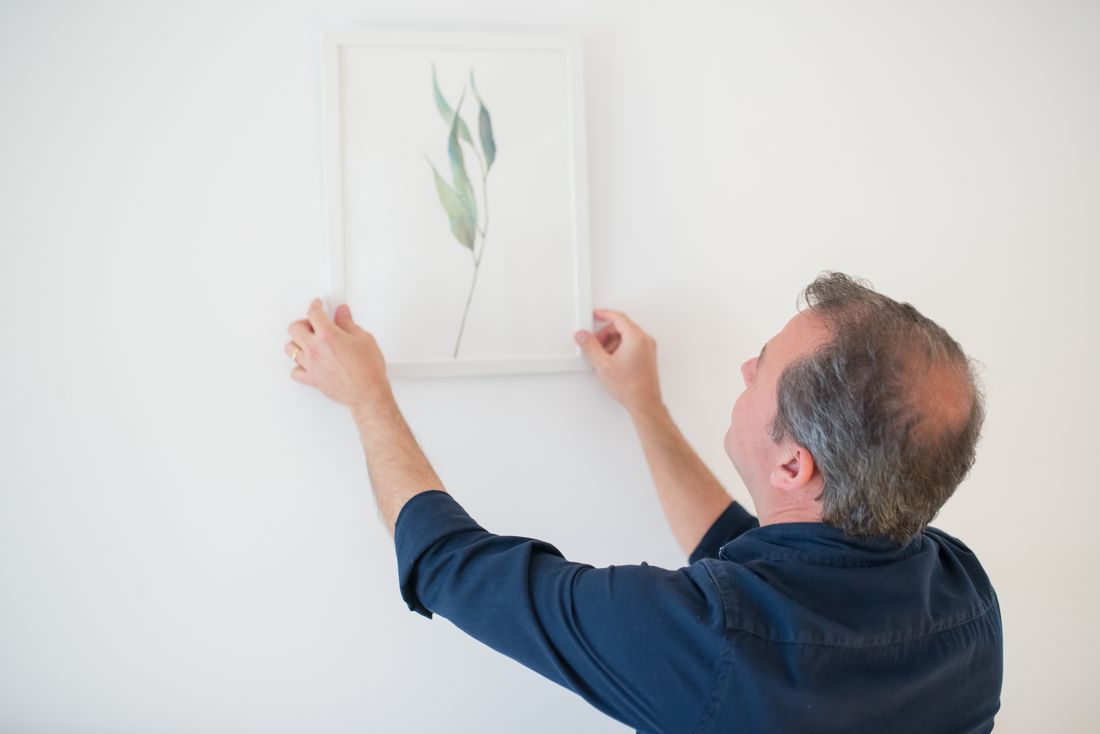 A man seen mostly from behind, reaching up with both hands to straighten a single framed botanical print on a plain white wall