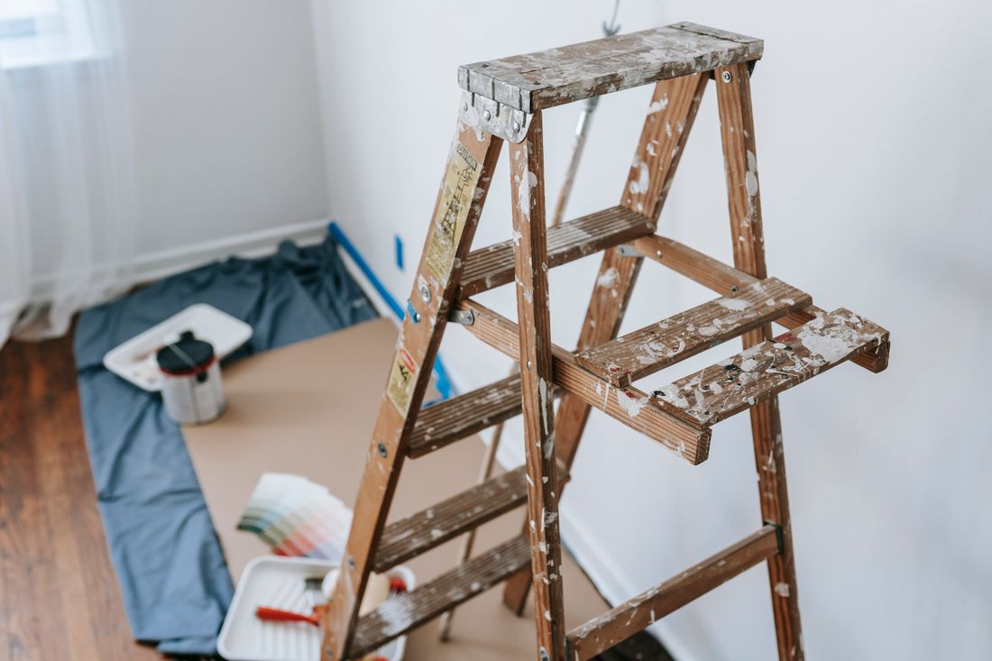 A paint-splattered wooden stepladder standing against a bare patch of wall, with a paint tray, color swatches, and drop cloths spread on the floor of a daylit room