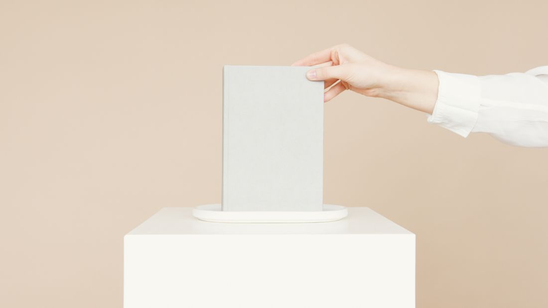 A hand steadying a plain book as it is set down on a low white pedestal against a neutral background