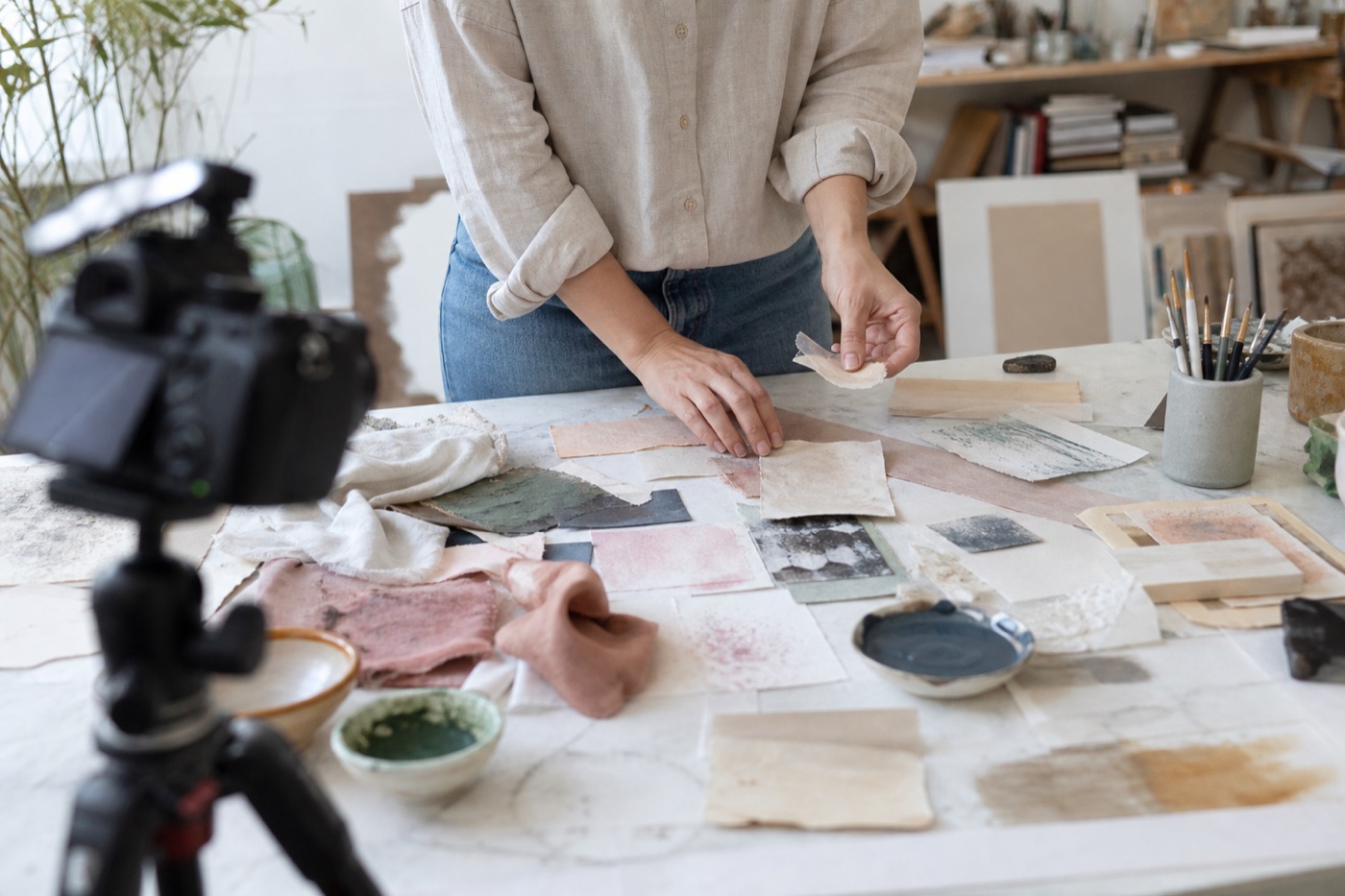 Artist arranging paper, fabric, pigment, and material samples while a camera records from a natural distance