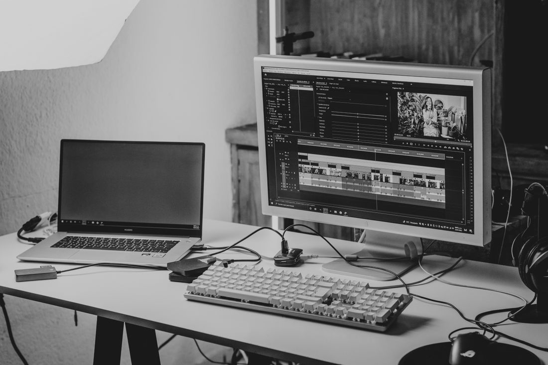 A monochrome home workspace with a laptop and an external monitor showing a video editing timeline
