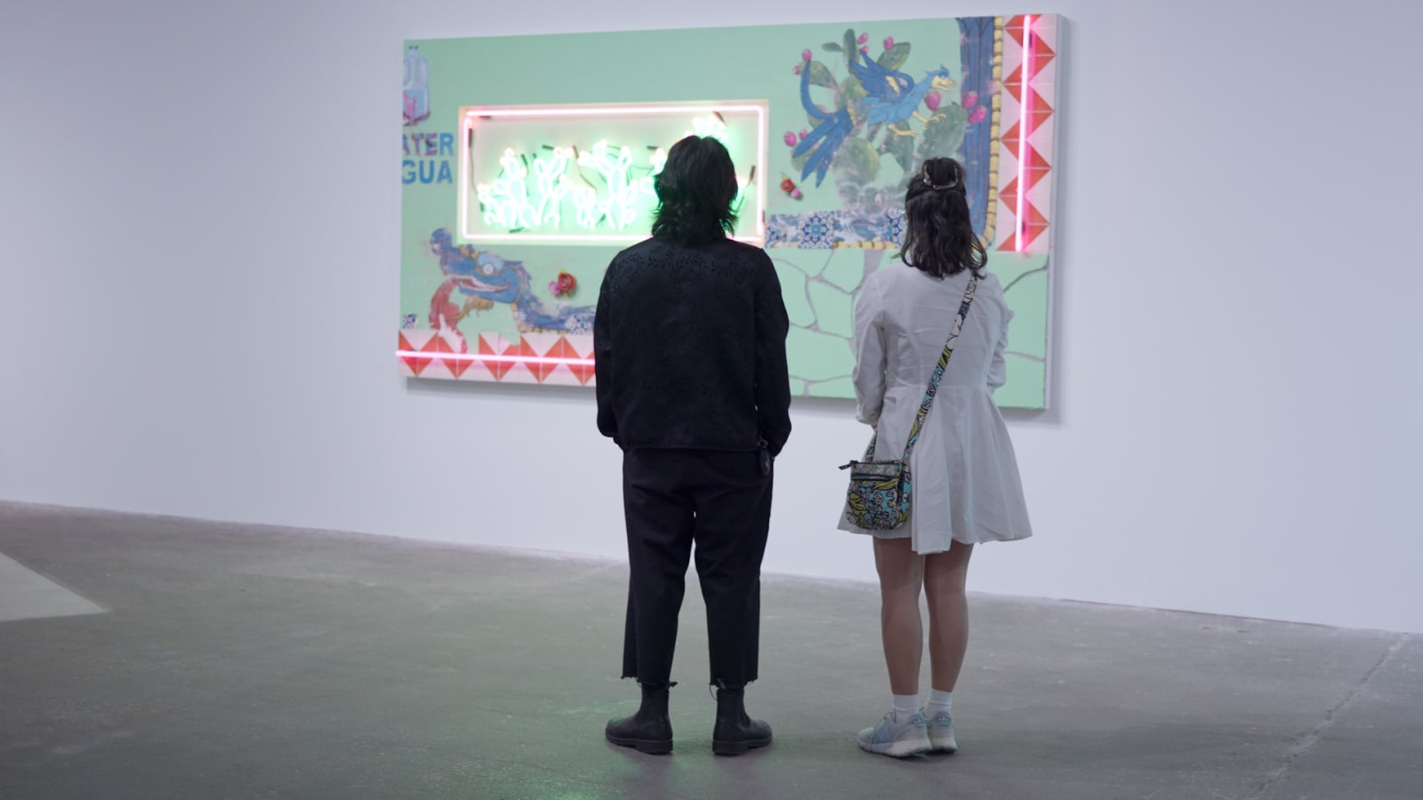 Two guests seen from behind studying a mixed-media wall piece with neon tubing and painted text