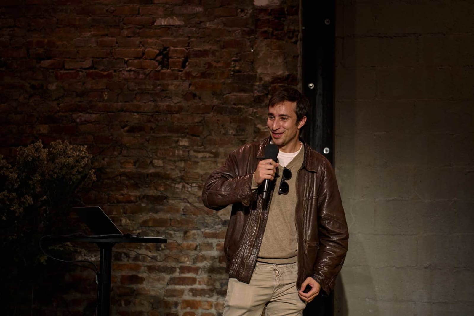 A speaker in a leather jacket addresses the room from in front of an exposed brick wall during an evening program