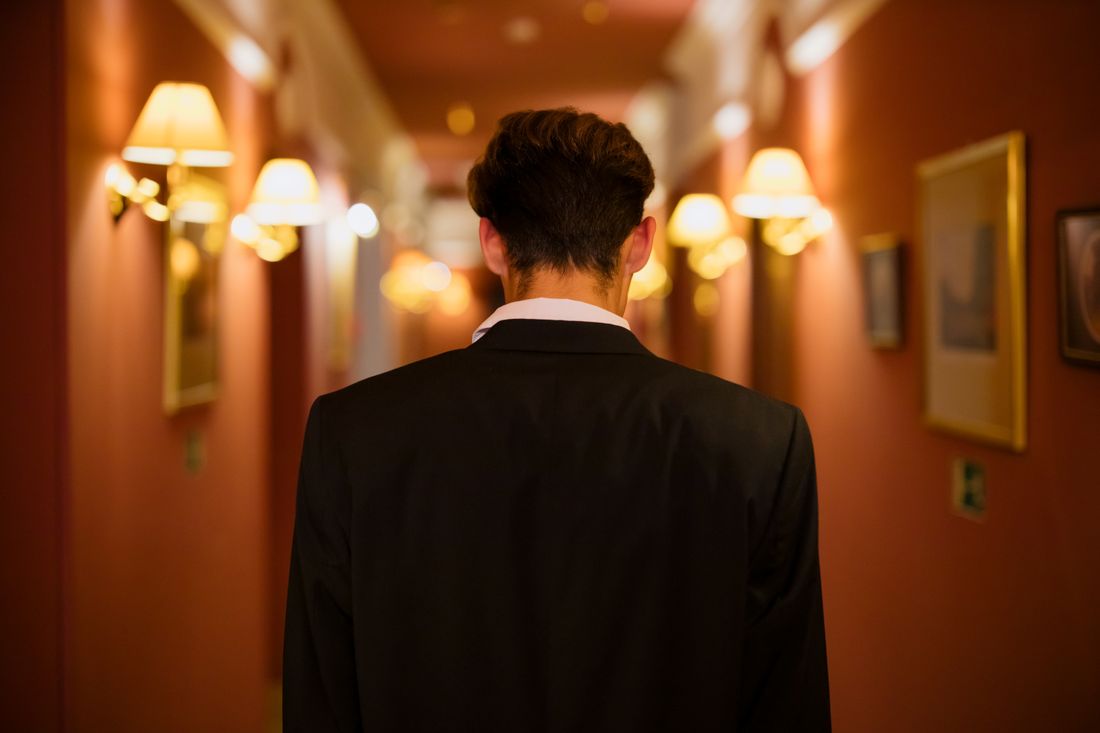 A person seen from behind, walking down a warmly lit corridor lined with wall sconces