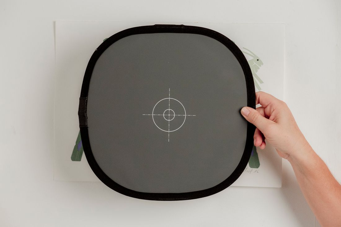 A hand holding a neutral gray target over a flat artwork during an imaging setup