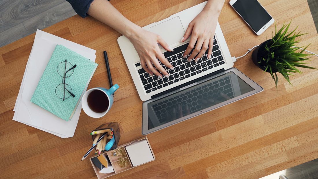 Overhead view of hands typing on a laptop at a wooden desk with coffee, a notebook, and a small plant