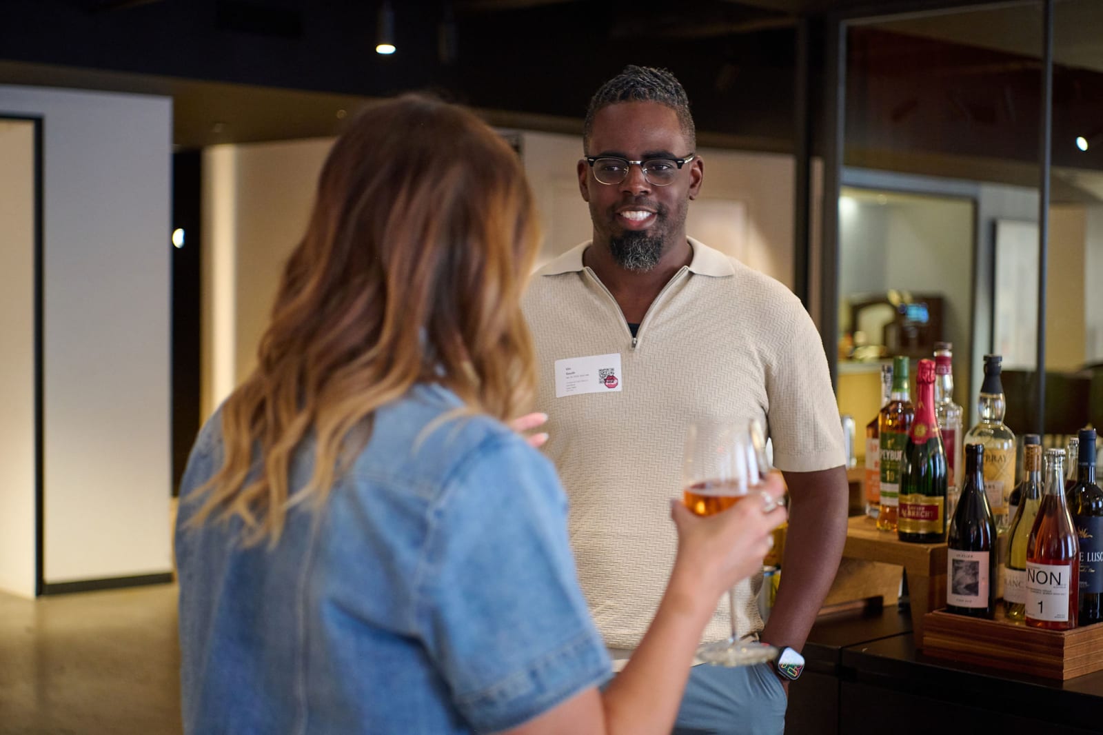 A gallery visitor wearing a name badge chats with another guest holding a glass of rose near a bar cart of bottles