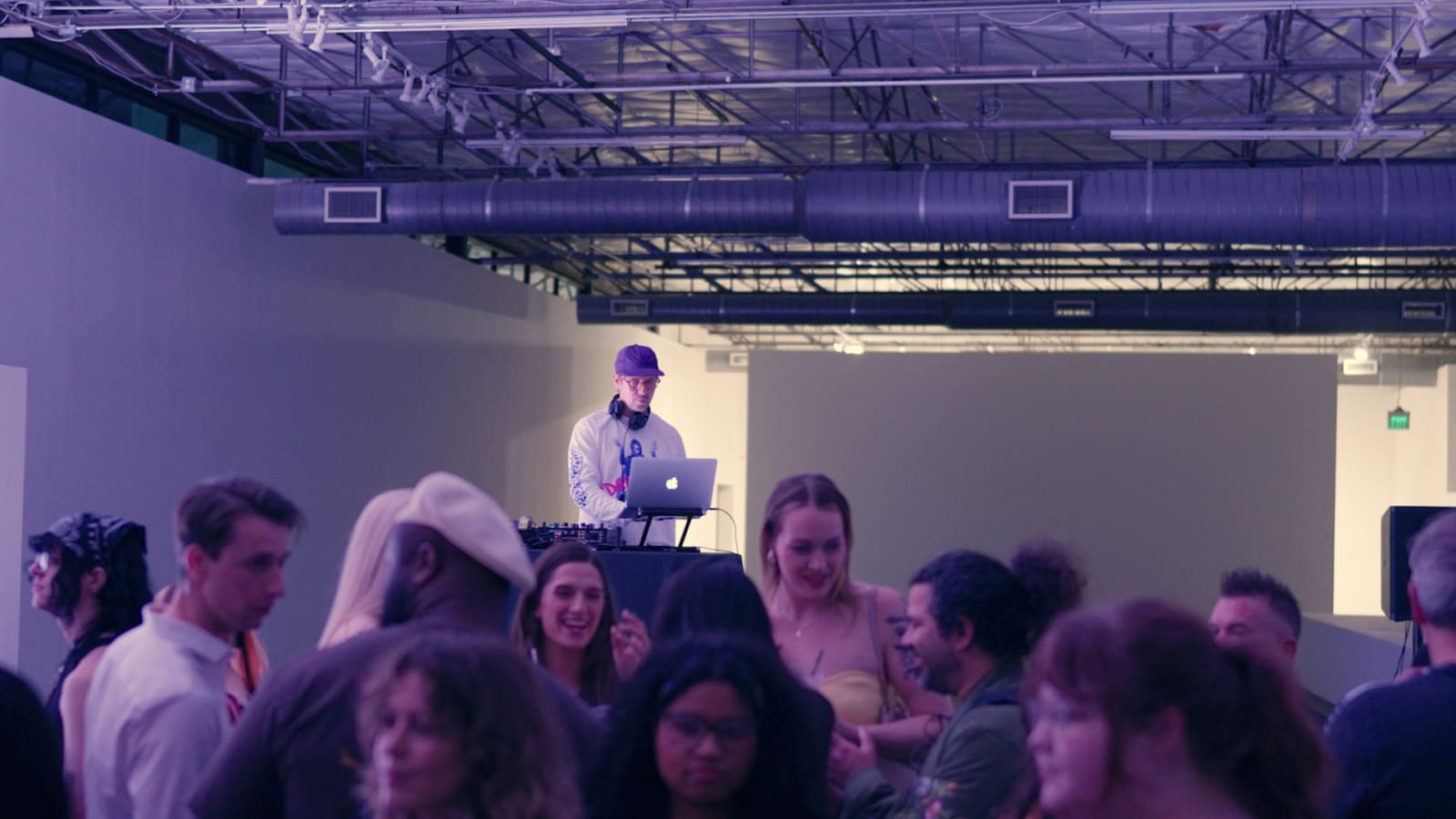 A DJ in a purple cap plays a laptop set behind a dense crowd dancing in a purple-lit gallery space