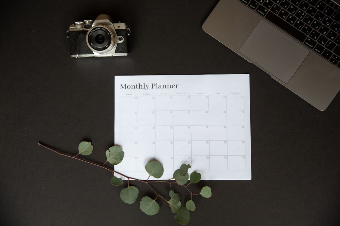 A monthly planner, camera, and laptop arranged on a desk for planning ahead