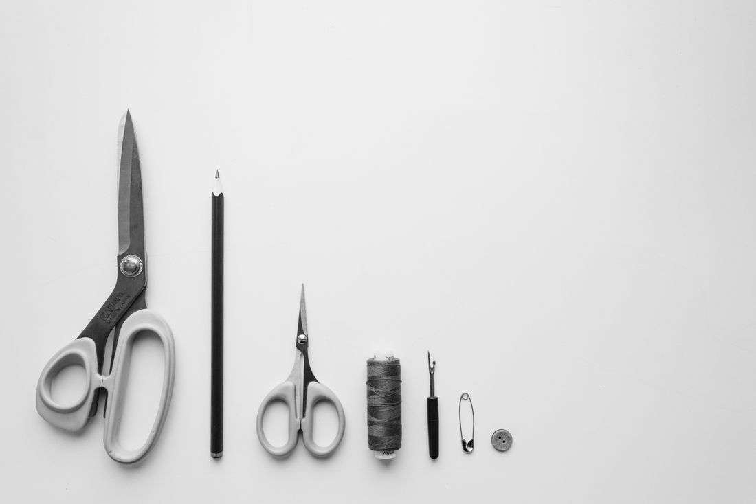 Minimalist flat lay of scissors, a pencil, thread, a needle, a safety pin, and a button on a white background