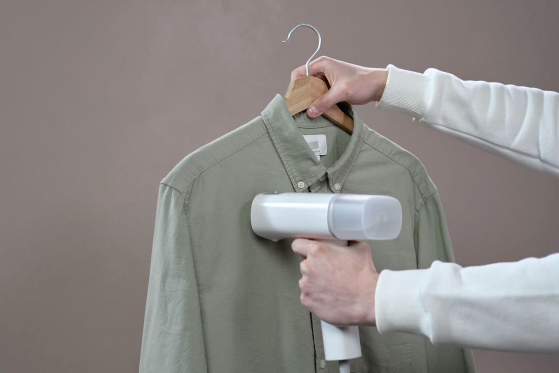Close crop of hands steaming a green shirt on a wooden hanger with a portable garment steamer