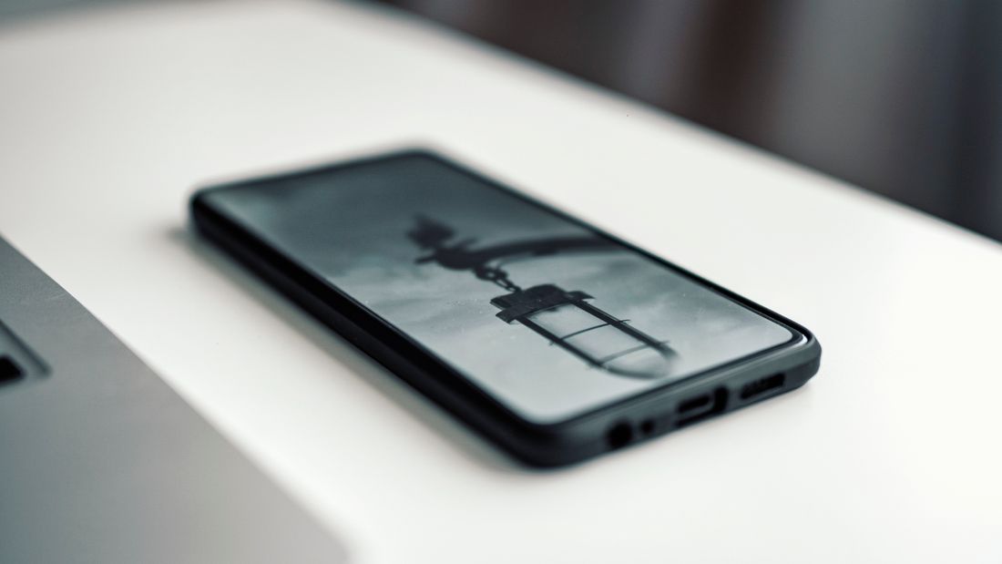A smartphone resting face-up on a white desk showing a dim, abstract paused video frame, screen content not legible, laptop edge visible nearby.