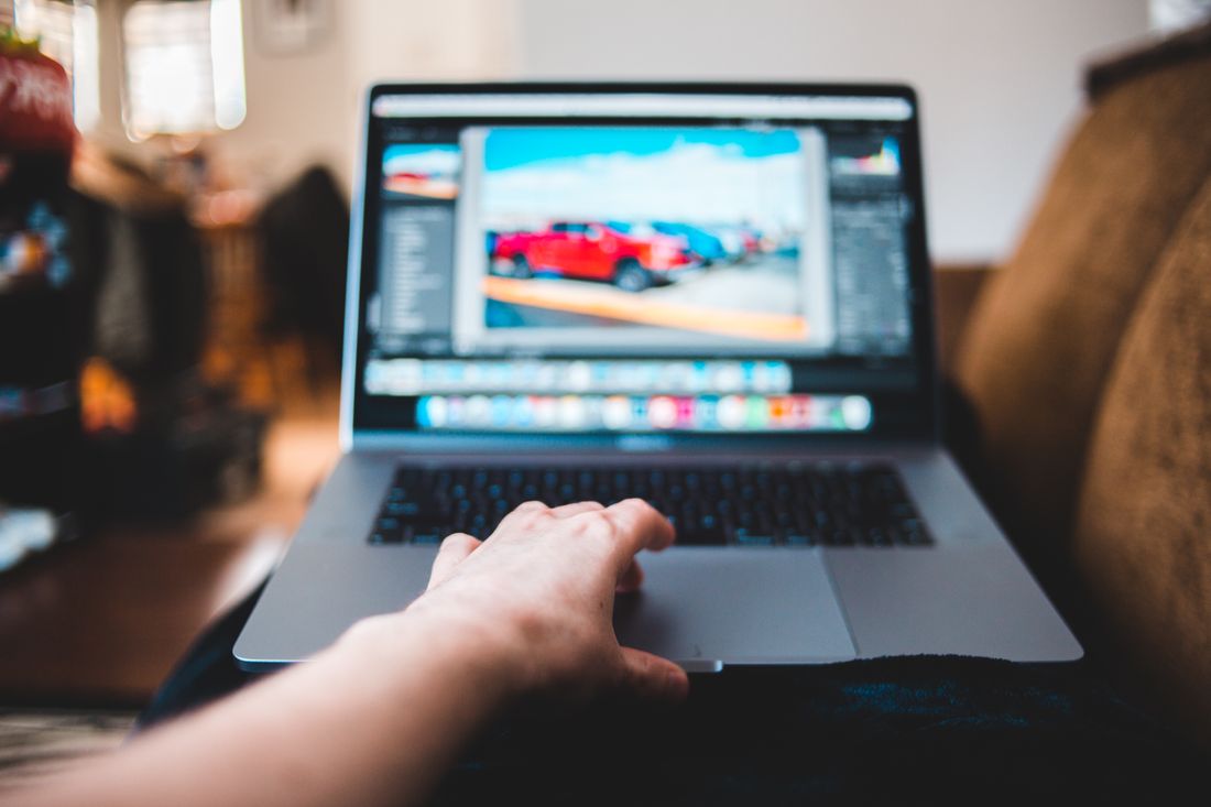 A hand reaching for the trackpad of an open laptop showing a soft-focus photo editing screen, shot from a low angle in a cozy living room.