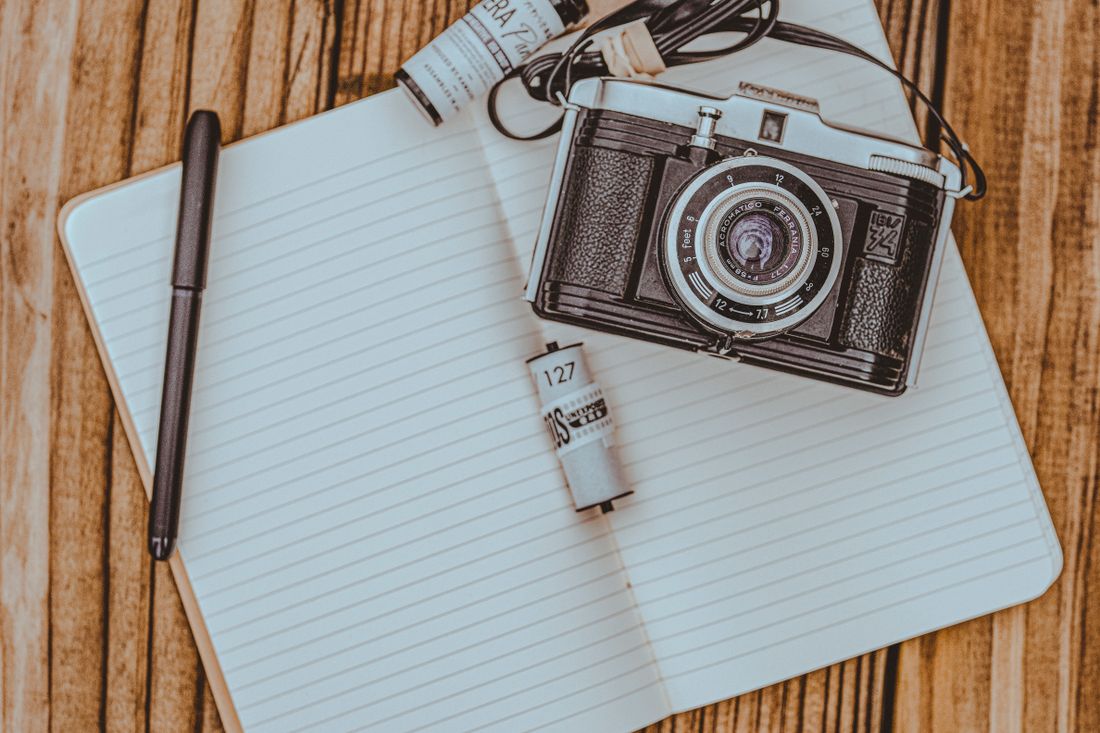 A small vintage camera and a roll of film resting on an open, blank notepad next to a pen, on a wooden table.