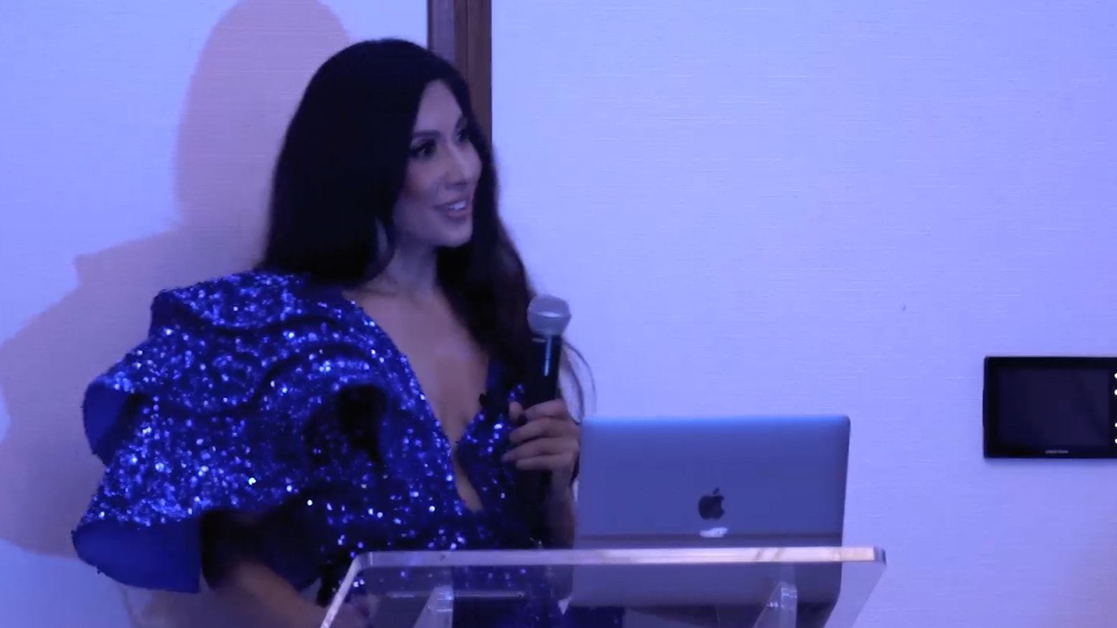 Speaker in a sequined dress addressing a room from a podium with a laptop and microphone under blue stage lighting